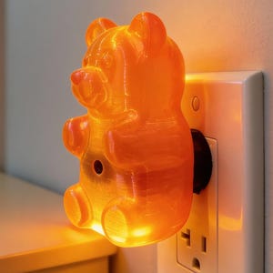 A warm orange gummy bear night light is plugged into a wall outlet in a child's bedroom, with a kid in the softly blurred background gazing happily at the illuminated bear. While it acts as a playful pop culture statue and a quirky housewarming gift for an apartment, this funky home accent plug in luminary is completely safe and generates no heat. It delivers a perfect kidcore aesthetic room vibe and serves as a highly rated dopamine decor gift, doubling as nostalgic neon polymer art decor.