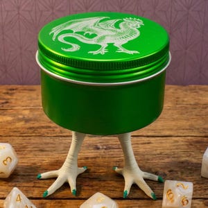 May include: A green container with a dragon design on the lid, supported by chicken feet. Several ivory-colored dice with gold numbering are scattered around the base, creating a whimsical and unique tabletop gaming accessory.