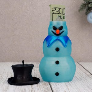 May include: A light blue snowman-shaped money bank with a menacing expression, an orange nose, and black buttons. A twenty-dollar bill is inserted into the top. A separate black top hat sits to the left of the snowman figure.