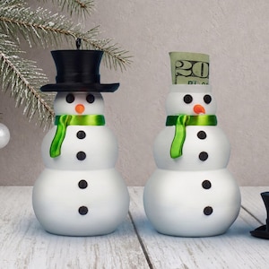 Two snowman ornaments side by side, one with its black top hat sealed on and one with the hat removed to reveal a rolled bill peeking from the hidden cash compartment inside its body.