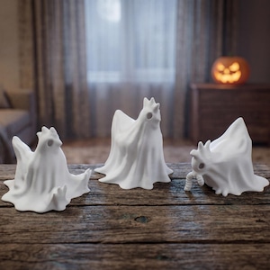 May include: Three white ceramic ghost figurines with draped cloaks and stylized faces sit on a rustic wooden table. The Halloween-themed decorations are arranged in a group, with a blurred background.