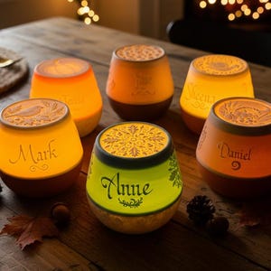 May include: Six illuminated personalized candle holders with names like "Mark" and "Anne" are arranged on a wooden table. The candle holders have a warm, orange glow and are decorated with intricate designs. Fall leaves and pinecones are scattered around.