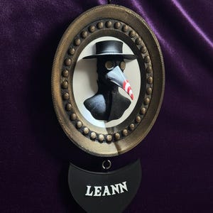 May include: A framed, oval-shaped wall hanging featuring a black plague doctor mask with a red and white striped beak. The frame is gold-colored with a beaded edge. Below the frame is a black, heart-shaped tag with the name "LEANN" in white.