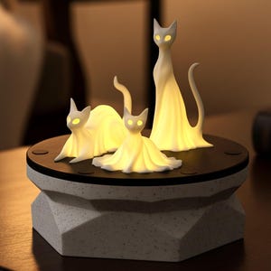 May include: A decorative cat lamp featuring three white cat figurines with glowing yellow eyes and draped forms. The cats are arranged on a black circular base atop a geometric, speckled, light gray pedestal. The lamp emits a warm, inviting glow.