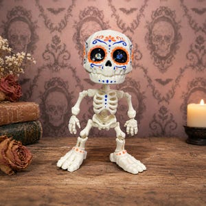 A close-up of the blue and orange patterned articulated skeleton standing proudly on a wooden dresser. This specific colorway perfectly captures the vibrant calavera art style, making this sugar skull figure a standout weirdcore accessory. Acting as an ADHD fidget tool for office or home, this tactile sensory item decor is the ultimate fun dopamine decor. A ready to ship goth gift and creepy cute pastel goth desk buddy for any alternative space.