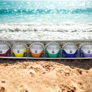 May include: A set of six colorful car-shaped ornaments in a box, each with a peace symbol. The cars are red, yellow, orange, green, blue, and purple, with white and silver accents. The background shows a sandy beach and ocean.