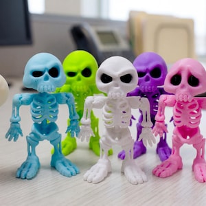 May include: A collection of five small, articulated skeleton figurines in vibrant colors including light blue, lime green, white, purple, and pink, each featuring a large skull with dark eye sockets and a detailed rib cage and jointed limbs.