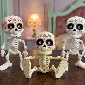 Three articulated skeleton toys displayed side by side in a cozy pastel goth bedroom setting. The center skeleton is sitting while the two outer figures stand, highlighting the posable nature of this ADHD fidget tool for office or home. A brilliant pastel goth vibe emanates from these vibrant calavera art pieces. An ideal witchy gift for friend or a quirky gift for adhd friend needing a fun dopamine decor element and weirdcore accessory.