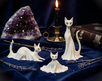 May include: Three white cat figurines with smooth, flowing, ghost-like silhouettes. One cat is standing upright, one is lying down, and one is a small, seated figure. Each cat has hollow, almond-shaped eye sockets and a minimalist, draped appearance.