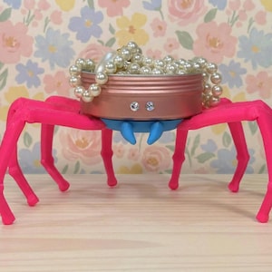 Puede incluir: Un objeto decorativo artesanal con forma de araña. Presenta patas de color rosa brillante que sostienen un recipiente color cobre lleno de perlas de imitación. La araña tiene pinzas azules y está adornada con pedrería.