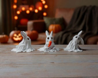 May include: Three white ceramic rabbit figurines, each draped in a ghost-like sheet. One rabbit holds a small orange carrot. The background includes pumpkins and a blurred string of lights, suggesting a Halloween theme.