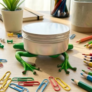 Może przedstawiać: Srebrny metalowy pojemnik z żebrowaną pokrywką spoczywa na zielonym uchwycie w kształcie żaby. Kolorowe ołówki, spinacze do papieru i pinezki otaczają pojemnik na drewnianej powierzchni. Widoczna jest również mała roślina doniczkowa i kubek.