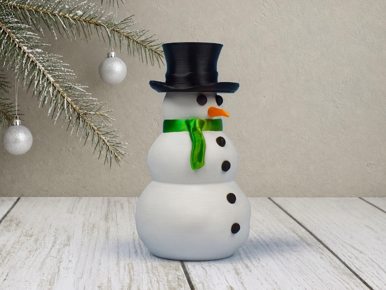 A snow-white snowman money holder ornament shown at an angle, its orange carrot nose and glossy green scarf bright against a beige plaster wall with a Christmas tree nearby, dopamine decor at its cheeriest.