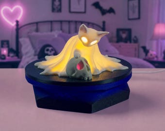 May include: A white cat-shaped lamp draped in a ghost-like sheet, emitting a warm yellow glow from its eyes and body. The lamp sits on a black circular base and rests next to a small, light-colored skull with a pink-tinted eye socket.