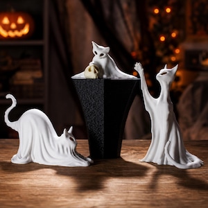 A complete set of the three cat ghost statues arranged on a rustic wooden surface, set against a background of festive Halloween decorations. The kitten sits on a central raised platform, with the stretching and reaching cats on either side, perfect as a whimsigoth Halloween decoration.