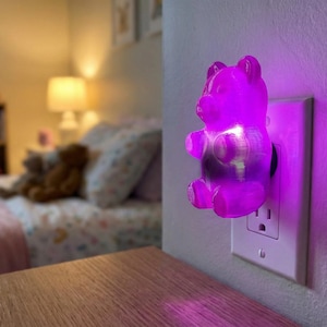 A brilliant purple gummy bear night light is plugged into a bedroom wall outlet, casting a soothing, colorful glow across the room's surfaces. This dopamine decor gummy bear night light serves as a delightfully eccentric fixture and a unique apartment gift for a friend. Functioning as a retro kitsch glowing candy art installation, this funky home accent plug in luminary instantly creates a weirdcore room vibe. It is the ideal choice when you want to buy weirdcore room accent piece bedroom decor.