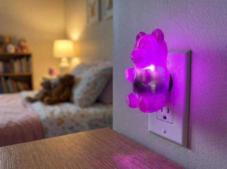 A brilliant purple gummy bear night light is plugged into a bedroom wall outlet, casting a soothing, colorful glow across the room's surfaces. This dopamine decor gummy bear night light serves as a delightfully eccentric fixture and a unique apartment gift for a friend. Functioning as a retro kitsch glowing candy art installation, this funky home accent plug in luminary instantly creates a weirdcore room vibe. It is the ideal choice when you want to buy weirdcore room accent piece bedroom decor.