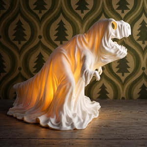 May include: A white dinosaur-shaped lamp draped in a sheet-like covering, resembling a ghost. The lamp emits a warm, soft orange glow from within, illuminating the textured folds of the material and highlighting the dinosaur's head and teeth.