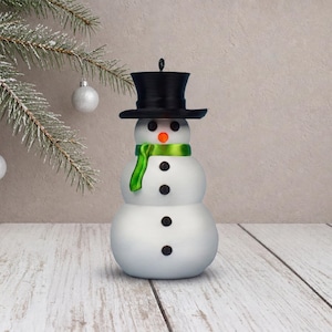 A classic snow-white snowman ornament with its black top hat, orange carrot nose, and lustrous green scarf, shown straight on against a beige plaster wall.