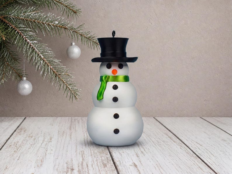 A classic snow-white snowman ornament with its black top hat, orange carrot nose, and lustrous green scarf, shown straight on against a beige plaster wall.