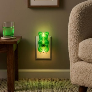 A bright green gummy bear night light is plugged into the wall of a warmly lit, cozy living room setting, adding a burst of vivid color to the relaxed environment. This maximalist gift operates as an eccentric fixture and a nostalgic ornament, providing the ultimate kidcore aesthetic room vibe. If you want to buy weirdcore room accent piece decor, this glowing candy shape is the perfect dopamine decor gummy bear night light to elevate your space with some retro kitsch glowing candy art magic.