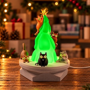 Pode incluir: Uma figura de árvore de Natal iluminada com uma estrela amarela no topo, apresentando cinco pequenas figuras de gatos nas cores laranja, castanho, preto e cinzento. Os gatos estão posicionados à volta e a subir a árvore verde, numa base branca.