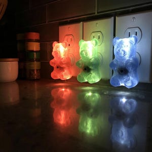 Three colorful gummy bear night light fixtures are plugged into a row of wall outlets situated just above a shiny, reflective kitchen countertop. This vibrant trio perfectly demonstrates how to use dopamine decor to brighten utilitarian spaces. Each glowing candy shape acts as an illuminated bear and a retro kitsch piece, making them a fantastic maximalist gift or quirky housewarming gift for an apartment. Transform your kitchen with this kidcore aesthetic funky home accent plug in luminary set.