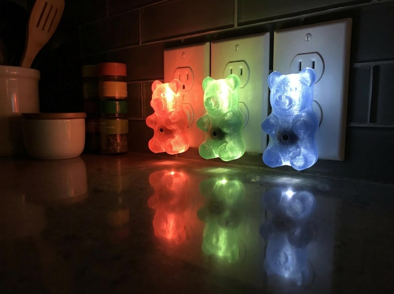 Three colorful gummy bear night light fixtures are plugged into a row of wall outlets situated just above a shiny, reflective kitchen countertop. This vibrant trio perfectly demonstrates how to use dopamine decor to brighten utilitarian spaces. Each glowing candy shape acts as an illuminated bear and a retro kitsch piece, making them a fantastic maximalist gift or quirky housewarming gift for an apartment. Transform your kitchen with this kidcore aesthetic funky home accent plug in luminary set.