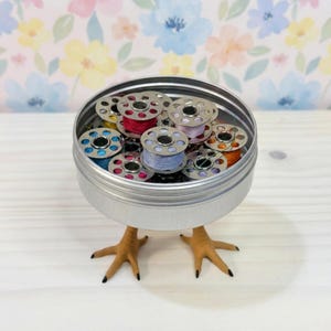 May include: A round silver metal tin filled with various colorful sewing machine bobbins, featuring light blue, pink, purple, and orange thread. The tin is supported by two realistic-looking tan bird feet attached to the base.