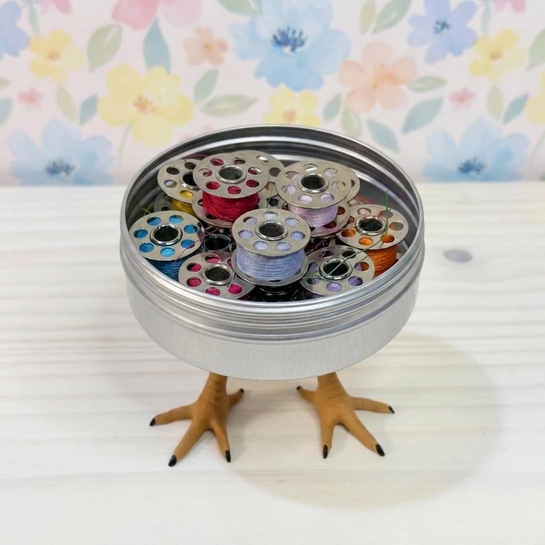 May include: A round silver metal tin filled with various colorful sewing machine bobbins, featuring light blue, pink, purple, and orange thread. The tin is supported by two realistic-looking tan bird feet attached to the base.