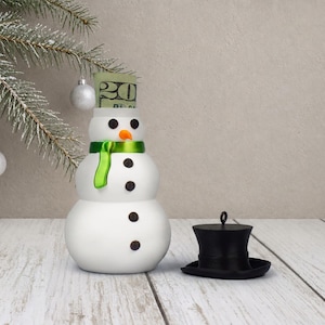 A single snowman money holder ornament with its black top hat lid set beside it and a rolled bill visible inside the hidden body compartment.