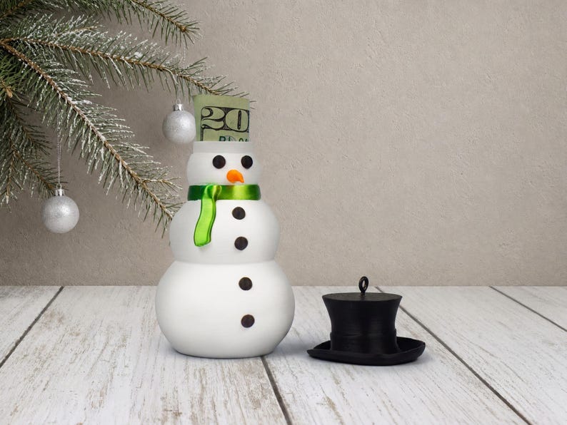 A single snowman money holder ornament with its black top hat lid set beside it and a rolled bill visible inside the hidden body compartment.