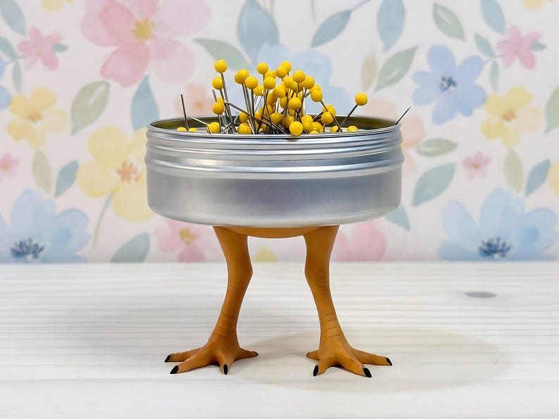 May include: A silver metal circular container filled with yellow-headed sewing pins, supported by two orange bird-like legs with black-tipped claws. The container is open at the top, revealing the collection of pins stored inside.