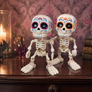 Two articulated skeletons standing side by side in a moody setting with a lit candle glowing softly in the background. These sugar skull figures serve as an evocative gothic home accent and a vibrant calavera art piece. Crafted as a tactile sensory item, this creepy cute pastel goth desk buddy is a fantastic witchy gift for friend. A wonderfully macabre weirdcore accessory and coworker gag gift that brings magic to any maximalist cubicle.