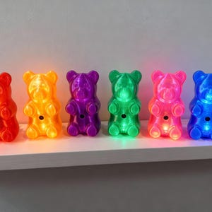 A glorious rainbow of six gummy bear night light luminaries lined up in a row on a wooden shelf, featuring vibrant shades of red, orange, purple, green, pink, and bright blue. Collection of dopamine decor showcases the ultimate retro kitsch piece options for anyone needing a quirky housewarming gift for an apartment. Each neon polymer art piece acts as a pop culture statue and a nostalgic ornament, allowing you to buy gummy bear night light sets to match your exact kidcore aesthetic room vibe.