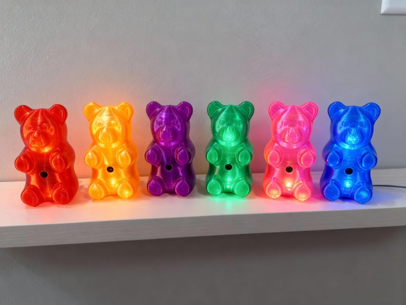 A glorious rainbow of six gummy bear night light luminaries lined up in a row on a wooden shelf, featuring vibrant shades of red, orange, purple, green, pink, and bright blue. Collection of dopamine decor showcases the ultimate retro kitsch piece options for anyone needing a quirky housewarming gift for an apartment. Each neon polymer art piece acts as a pop culture statue and a nostalgic ornament, allowing you to buy gummy bear night light sets to match your exact kidcore aesthetic room vibe.