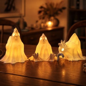 Op de afbeelding: Drie oplichtende spookvormige decoraties op een houten tafel. De witte figuren stralen een warm, geel licht uit en hebben kleine, ronde ogen. Eén spook heeft een worm. De scène speelt zich af in een gezellige kamer.