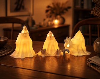 May include: Three glowing ghost-shaped decorative lights on a wooden table. The lights are white with a soft, yellow glow, and feature small, round eyes. One ghost has a worm. The scene is set in a warm, inviting room.