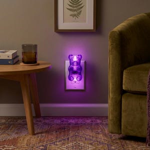 A radiant purple gummy bear night light is plugged into a cozy living room wall outlet, hovering just above a plush carpet with a comfortable couch resting nearby in the background. This eccentric fixture provides a weirdcore room vibe, making it the perfect dopamine decor gift for adults. The glowing candy shape serves as an illuminated bear and a funky home accent plug in luminary, offering a unique plug in outlet light solution for anyone wanting to buy a retro pop art maximalist gift.