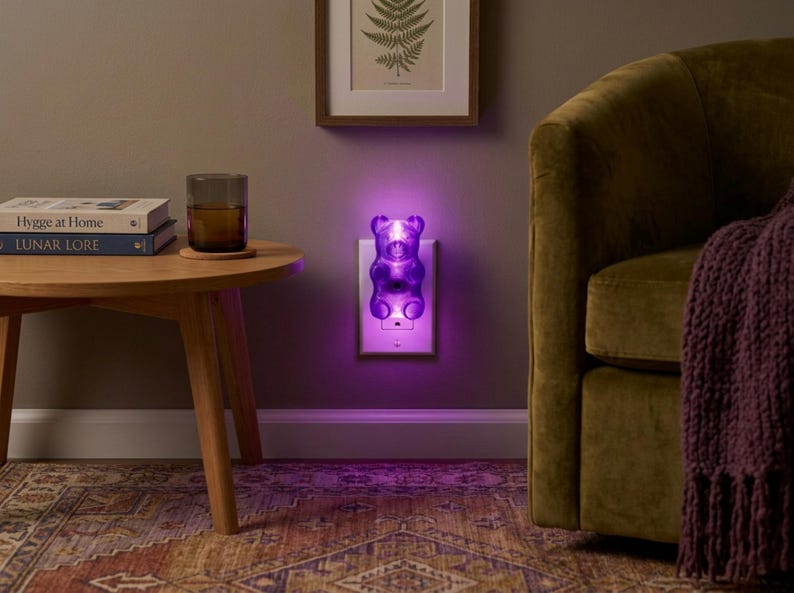 A radiant purple gummy bear night light is plugged into a cozy living room wall outlet, hovering just above a plush carpet with a comfortable couch resting nearby in the background. This eccentric fixture provides a weirdcore room vibe, making it the perfect dopamine decor gift for adults. The glowing candy shape serves as an illuminated bear and a funky home accent plug in luminary, offering a unique plug in outlet light solution for anyone wanting to buy a retro pop art maximalist gift.