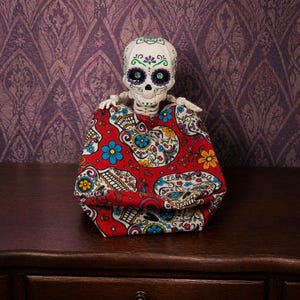 An articulated skeleton tucked comfortably inside a bright red, sugar skull themed fidget bag, resembling a cozy sleeping bag. This optional accessory enhances the weirdcore desk fidget toy experience, providing extra texture for this gothic tactile sensory item decor. It transforms the creepy cute pastel goth desk buddy into an exceptional quirky gift for adhd friend or a unique coworker gag gift. A delightful piece of fun dopamine decor.