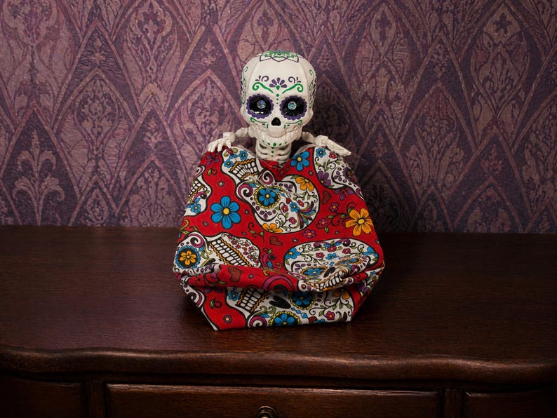 An articulated skeleton tucked comfortably inside a bright red, sugar skull themed fidget bag, resembling a cozy sleeping bag. This optional accessory enhances the weirdcore desk fidget toy experience, providing extra texture for this gothic tactile sensory item decor. It transforms the creepy cute pastel goth desk buddy into an exceptional quirky gift for adhd friend or a unique coworker gag gift. A delightful piece of fun dopamine decor.