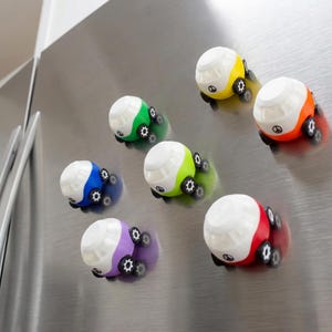 Op de afbeelding: Een verzameling kleurrijke speelgoedautomagneten in verschillende kleuren, waaronder geel, groen, oranje, rood, blauw en paars. Elke auto heeft een wit dak en zwarte wielen, bevestigd op een roestvrijstalen oppervlak.