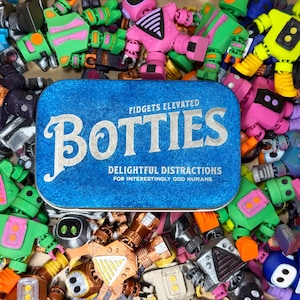 May include: A blue tin with the word "BOTTIES" in white lettering, surrounded by colorful robot-like toys. The tin reads "Fidgets Elevated" and "Delightful Distractions for Interestingly Odd Humans."