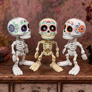 A trio of articulated skeletons where the center figure looks directly at the camera while the other two gaze inward. This image perfectly highlights the contrast between the natural bone and pure white color options of this sugar skull figure. An excellent gothic home accent and weirdcore desk fidget toy. This creepy cute pastel goth desk buddy is an engaging tactile sensory item, serving as a unique coworker gag gift and fun dopamine decor.