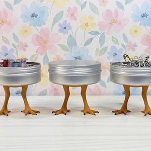 May include: Three round silver metal tins, each supported by a pair of yellow-orange bird-like legs with three-toed feet. The left tin contains colorful thread spools, the center tin is closed, and the right tin holds silver safety pins.