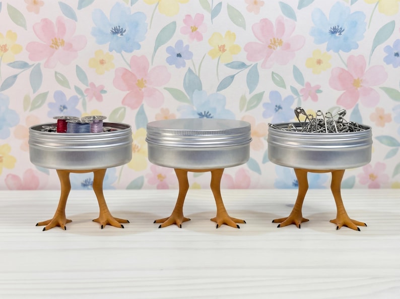 May include: Three round silver metal tins, each supported by a pair of yellow-orange bird-like legs with three-toed feet. The left tin contains colorful thread spools, the center tin is closed, and the right tin holds silver safety pins.