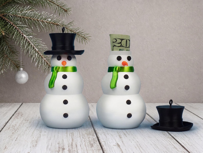 Two snowman ornaments side by side, one with its black top hat sealed on and one with the hat removed to reveal a rolled bill peeking from the hidden cash compartment inside its body.