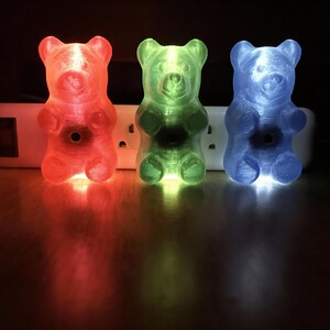 Puede incluir: Tres luces de noche con forma de osito de gominola en rojo, verde y azul. Las luces con forma de oso est&aacute;n enchufadas a una regleta blanca. Las luces proyectan un brillo colorido sobre la superficie.