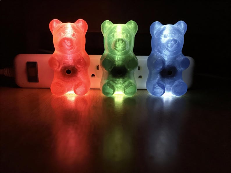 Puede incluir: Tres luces de noche con forma de osito de gominola en rojo, verde y azul. Las luces con forma de oso est&aacute;n enchufadas a una regleta blanca. Las luces proyectan un brillo colorido sobre la superficie.